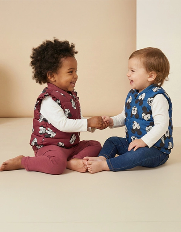 Dvě batolata sedí před světlým pozadím, mají na sobě barevné vesty, drží se za ruce a usmívají se na sebe.