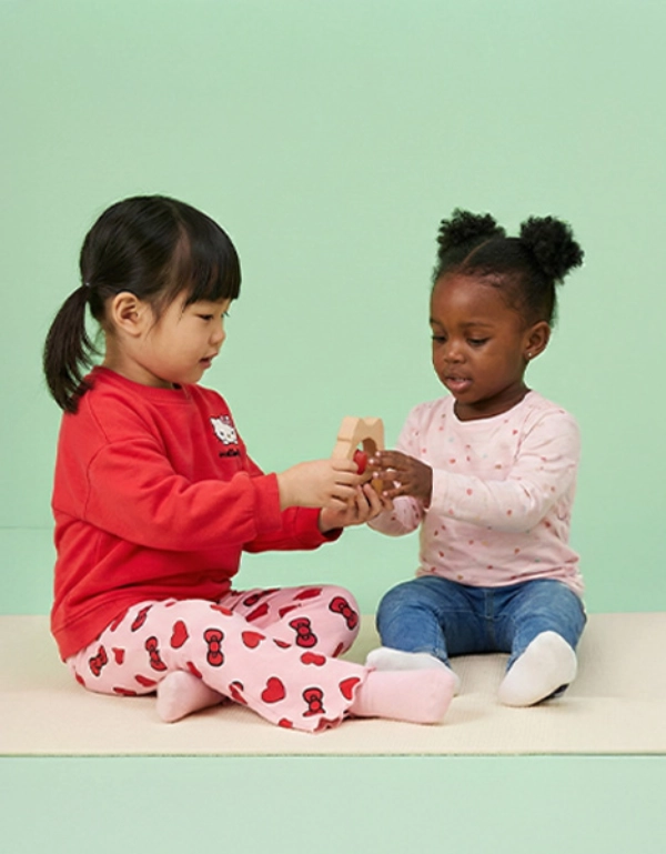 Dvě malé děti sedí na měkké podložce a radostně si spolu hrají, zatímco drží barevnou hračku před pastelově zeleným pozadím.