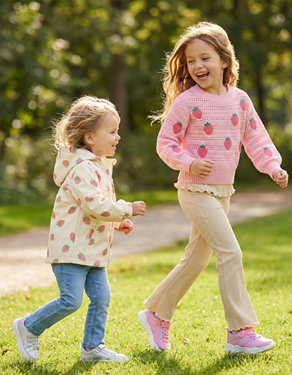 Dvě děti vesele běhají po sluncem zalitém parku obklopeném zelenou trávou a stromy a mají na sobě barevné svetry a volnočasové kalhoty.
