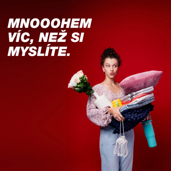 Osoba drží několik barevných předmětů, včetně přikrývek, láhve s vodou a květin, na pozadí zářivě červené barvy.