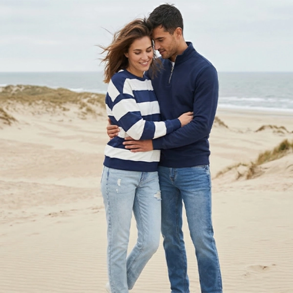 Pár v ležérním oblečení se objímá na písečné pláži s dunami a mořskými vlnami pod zataženou oblohou.