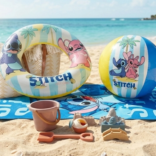 Plážová osuška, nafukovací kruh, míč a hračky na písek s motivem Lilo a Stitch leží na písečné pláži u tyrkysového moře.