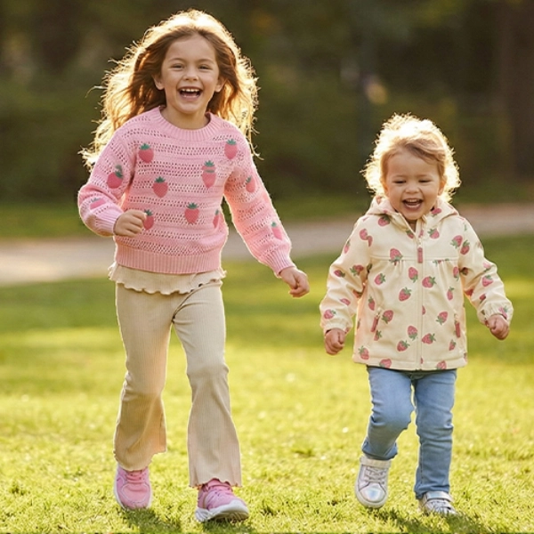 Dvě děti radostně běží sluncem zalitým parkem, obklopené zelenou trávou a stromy, oblečené v barevných svetrech a ležérních kalhotách.