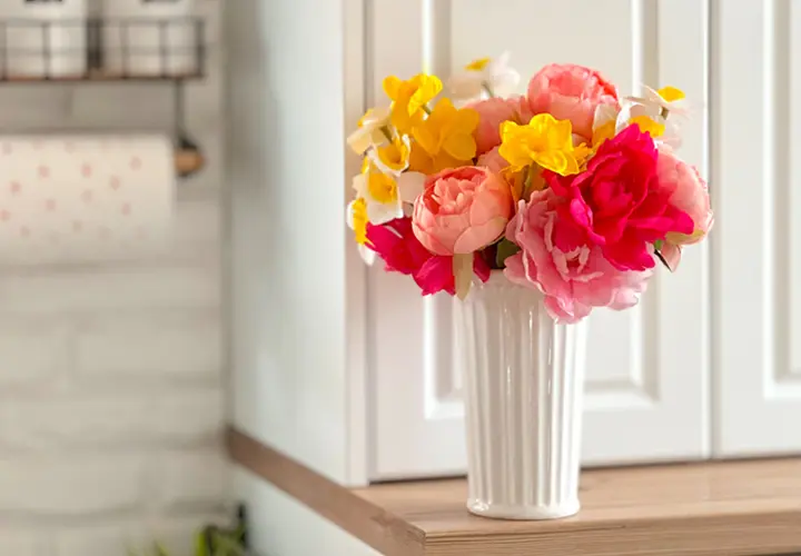 Barevné umělé květiny v bílé váze vystavené na dřevěné polici na světlém pozadí kuchyně.