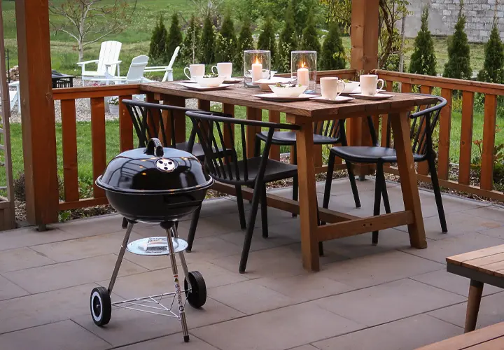 Útulné venkovní posezení s dřevěným stolem se svíčkami, židlemi a malým černým grilem vedle, uprostřed zelené zahrady.