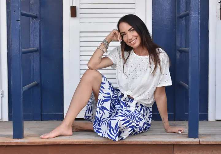 Na verandě sedí půvabná žena v bílém topu a modré květované sukni, ruku má položenou na bradě a je hluboce zamyšlená.