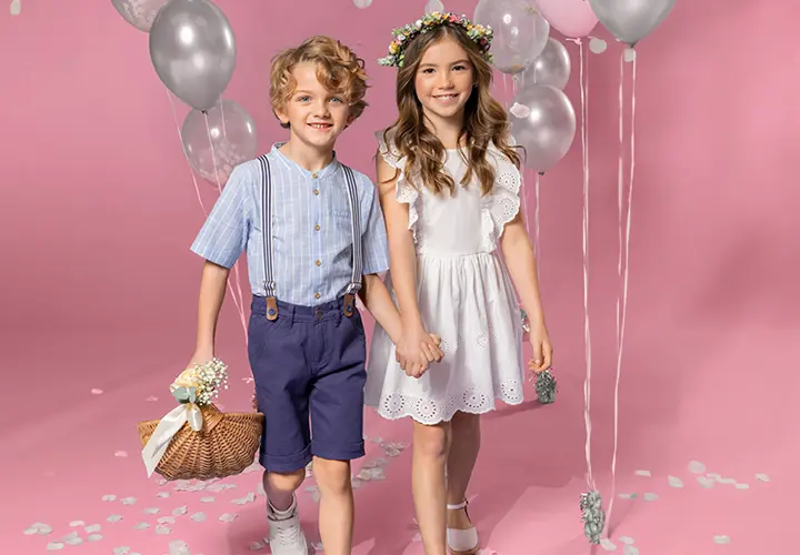 Dvě děti se drží za ruce a stojí na růžovém pozadí. Jedno má na sobě pruhované tričko a šortky, druhé bílé šaty a květinovou korunku.