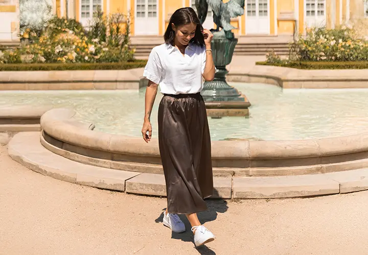 Žena v bílém tričku a hnědé sukni kráčí u fontány obklopené zelení, v pozadí žluté budovy.