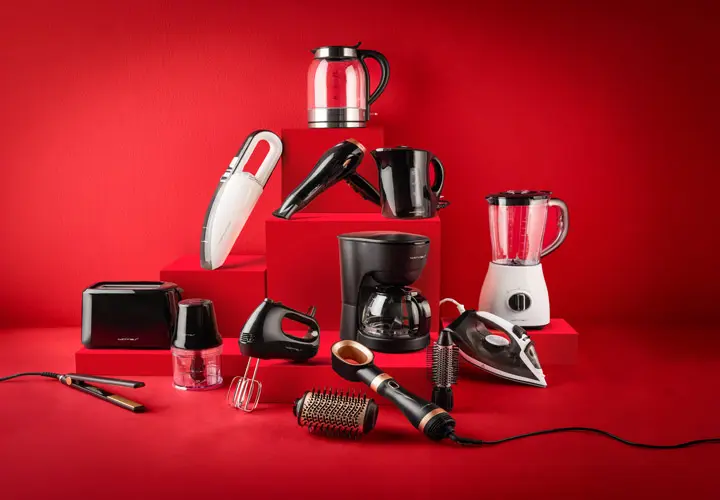 Sbírka různých domácích spotřebičů, včetně konvice, mixéru, kávovaru a vysavače, uspořádaných na červených schodech.