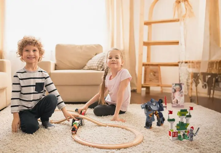 Dvě děti si hrají na kobercové podlaze s dřevěnou vláčkodráhou, hračkovým robotem a stavebnicí, obklopené útulným obývacím pokojem.