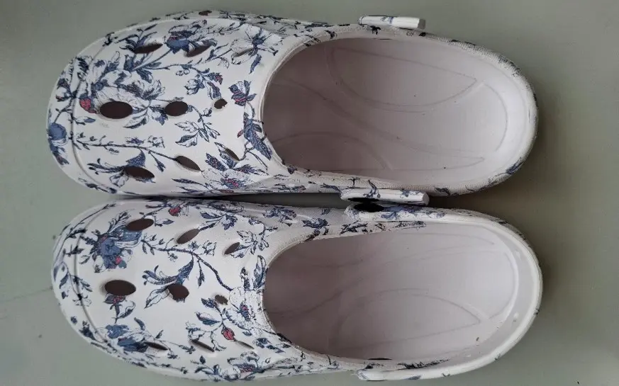 Pár bílých dřeváků s květinovým vzorem a ventilačními otvory, pohled shora, položené na rovném povrchu. Pár bílých dřeváků s květinovým vzorem a ventilačními otvory, pohled shora, položené na rovném povrchu.