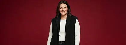 Osoba stojí sebevědomě před jasně červeným pozadím, oblečená v bílém tričku s dlouhým rukávem a černé vestě.