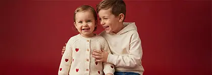 Dvě děti pózují těsně vedle sebe, jedno v měkkém svetru se srdíčky a druhé ve světlé mikině s kapucí, před červeným pozadím.