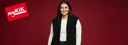 Osoba stojí sebevědomě před jasně červeným pozadím, oblečená v bílém tričku s dlouhým rukávem a černé vestě.