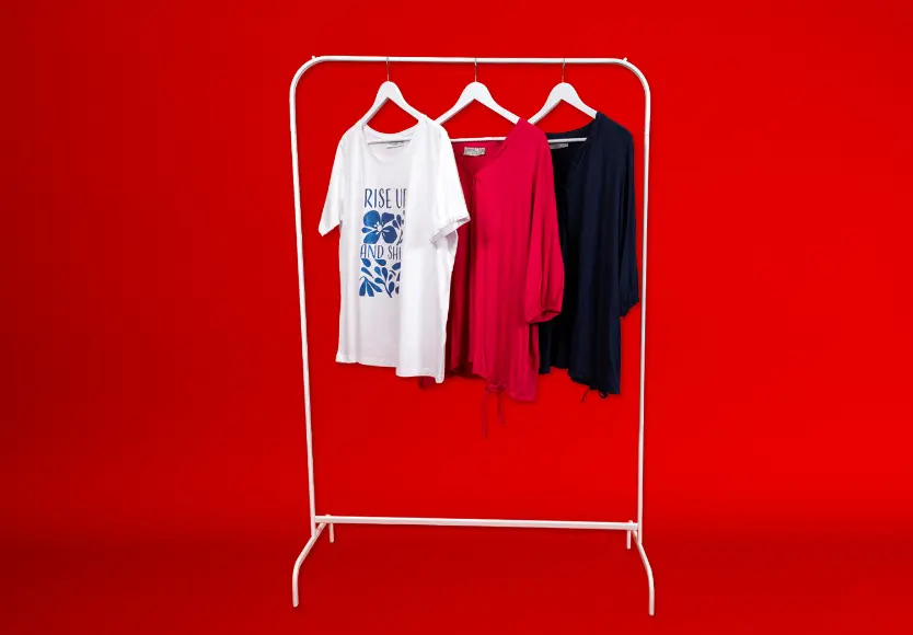 Tři stylové vrchní díly visí na bílém stojanu před sytě červeným pozadím: bílé tričko s potiskem, růžová halenka a námořnicky modrá košile.