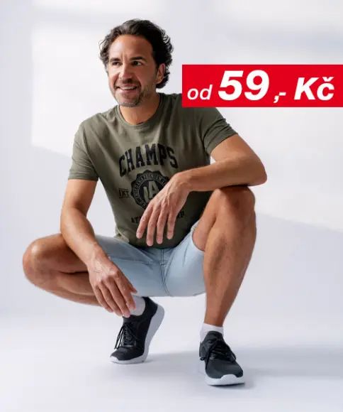 Muž v ležérní póze, oblečený do zeleného trička a světle modrých šortek, s černými sportovními botami, před jemným pozadím.