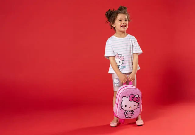 Dítě stojí před červeným pozadím, má na sobě pruhované tričko a kraťasy s motivem Hello Kitty a drží růžový batoh Hello Kitty