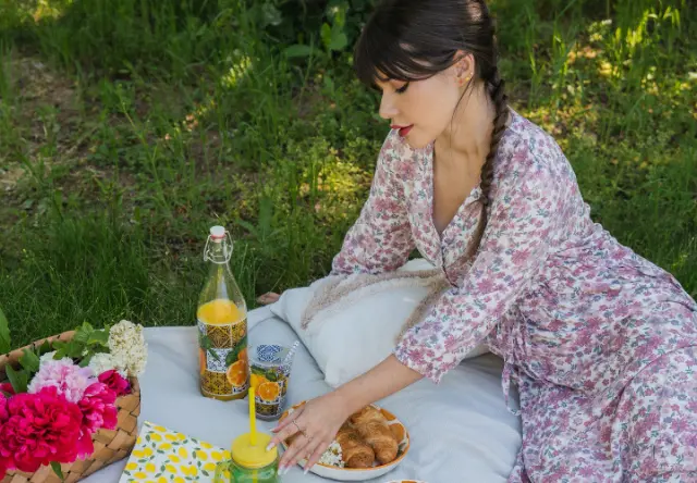 Scéna z pikniku, na které je žena v květinových šatech obklopená zářivými květinami, pomerančovým nápojem, croissanty a barevnou dekou na trávě.