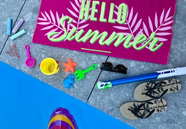 Živá letní scéna s růžovým ručníkem s nápisem „Hello Summer“, plážovými hračkami, slunečními brýlemi a žabkami vedle bazénu.