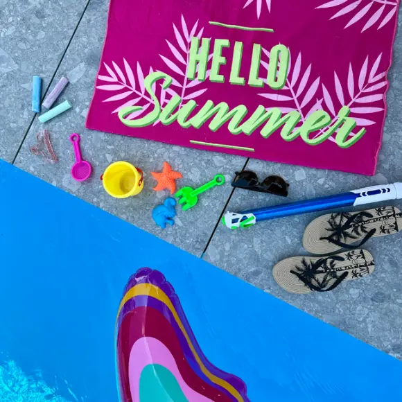Živá letní scéna s růžovým ručníkem s nápisem „Hello Summer“, plážovými hračkami, slunečními brýlemi a žabkami vedle bazénu.