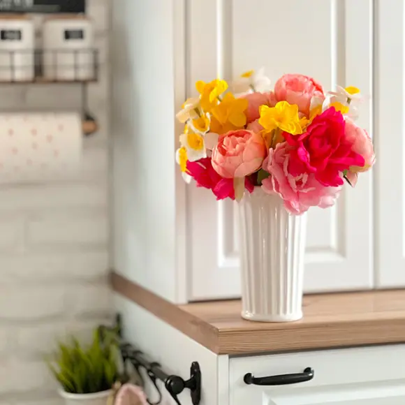 Barevné umělé květiny v bílé váze vystavené na dřevěné polici na světlém pozadí kuchyně.