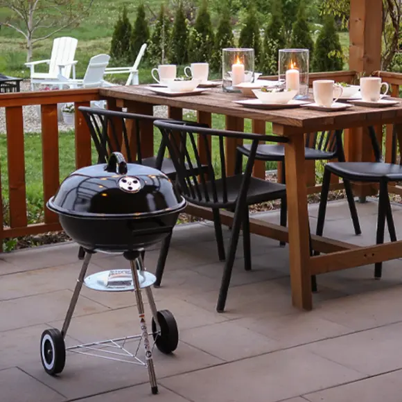 Útulné venkovní posezení s dřevěným stolem se svíčkami, židlemi a malým černým grilem vedle, uprostřed zelené zahrady.