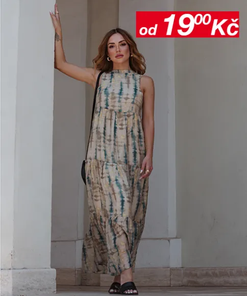 Žena v batikovaných maxi šatech stojí u zdi. V rohu je vidět cedule s cenami.