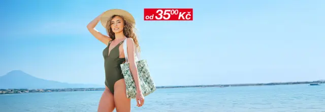 Žena v zelených plavkách a klobouku se širokou krempou stojí na pláži a drží tašku s tropickým motivem pod jasnou oblohou.