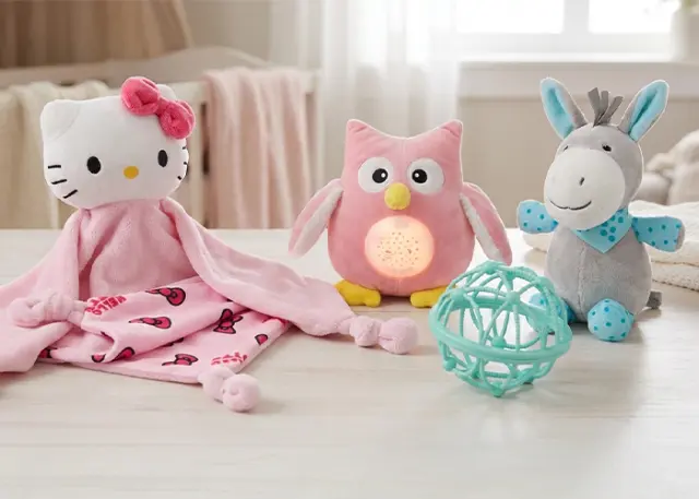 Útulné uspořádání plyšových hraček: deka s Hello Kitty, růžová sova se světýlkem, šedý oslík a tyrkysová koule, vše na dřevěném stole.