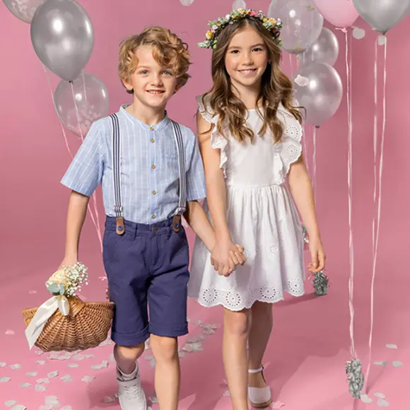 Dvě děti se drží za ruce a stojí na růžovém pozadí. Jedno má na sobě pruhované tričko a šortky, druhé bílé šaty a květinovou korunku.