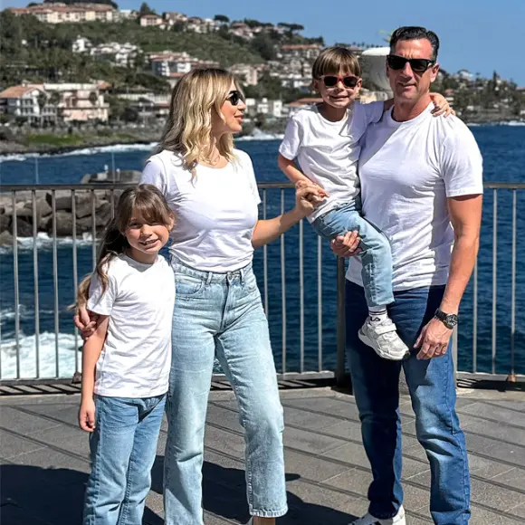 Čtyřčlenná rodina pózuje společně u moře, oblečená do ladících bílých triček a džínů, s malebnou kulisou kopců a vody v pozadí.