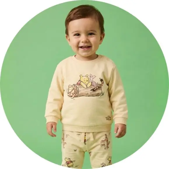 Batole stojí před zeleným pozadím a má na sobě žlutý svetr s hravou ilustrací kreslených postaviček.