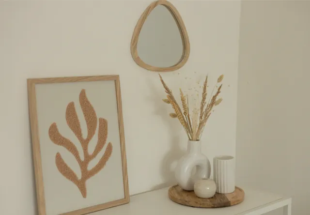 Minimalistický dekor s malým zrcadlem, zarámovaným listovým uměním a vázou se sušenými květinami na dřevěném podnosu, na pozadí neutrální stěny.