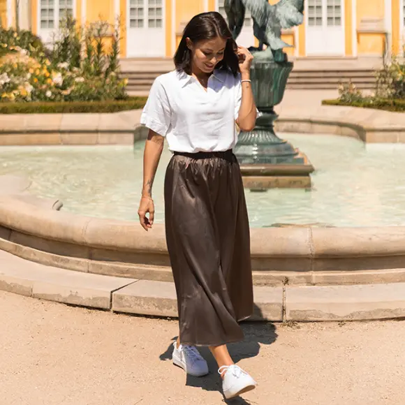 Žena v bílém tričku a hnědé sukni kráčí u fontány obklopené zelení, v pozadí žluté budovy.