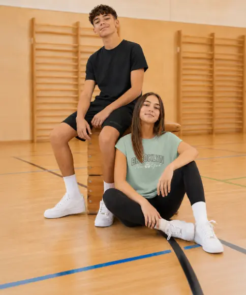 Dva teenageři v pohodlném sportovním oblečení sedí v tělocvičně na přeskakovací bedně a vedle ní. Oblečení je ideální pro školní tělocvik.