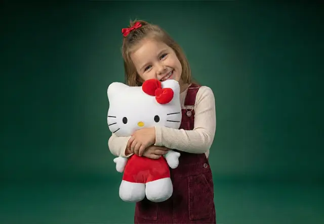Dítě v bordó outfitu objímá velkou plyšovou hračku Hello Kitty na zeleném pozadí.