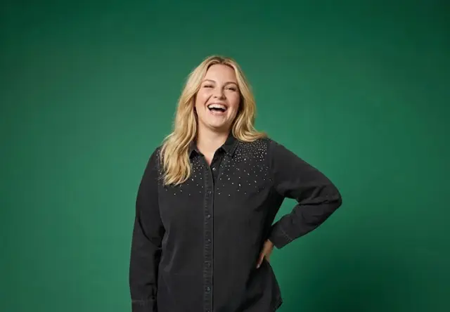 Žena s vlnitými blond vlasy stojí sebevědomě v černém tričku s jemnými ozdobnými detaily na zeleném pozadí.