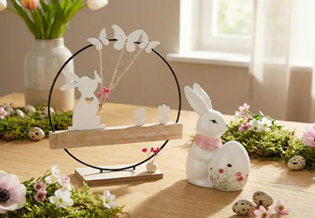 Dekorativní jarní scéna s figurkou zajíčka a vajíčkem, obklopená motýly, květinami a zelení na dřevěném stole.