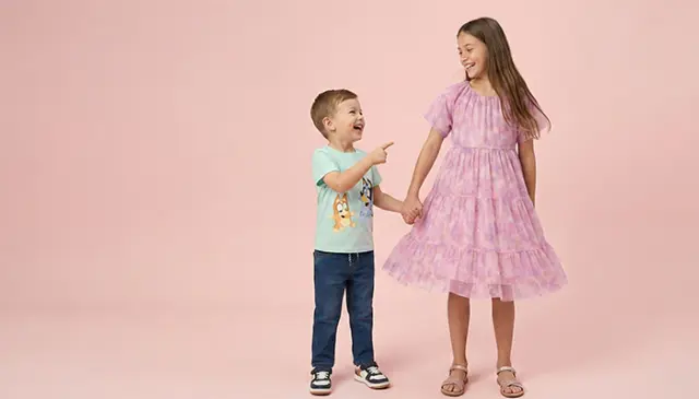 Mladý chlapec v světle modré košili a džínách ukazuje na dívku v růžových šatech s volánky, oba se drží za ruce na pozadí jemně růžové barvy.