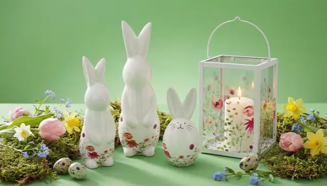Tři keramické zajíčky, vzorované vejce a lucerna se svíčkou, obklopené barevnými květinami a velikonočními vajíčky na zeleném pozadí.