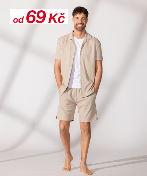 Muž v ležérní póze, oblečený do zeleného trička a světle modrých šortek, s černými sportovními botami, před jemným pozadím.
